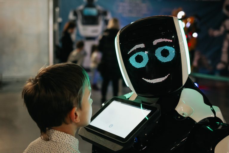 A young boy is looking at a futuristic robot. There is a tablet in front of the machine. The robot is smiling at the boy, who is looking up in wonder.