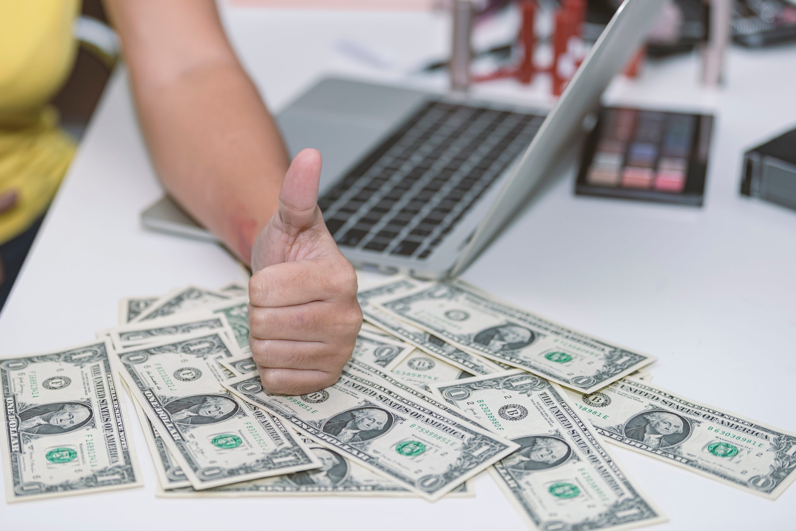 Dollars scattered over a table with someone making a thumbs-up sign on top of them, in front of their laptop.