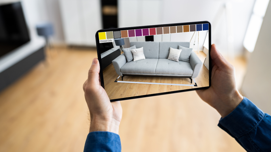 Someone holds a tablet in their living room. On the screen of the tablet, there is an image of a large, grey sofa. A selection of colours run across the top.
