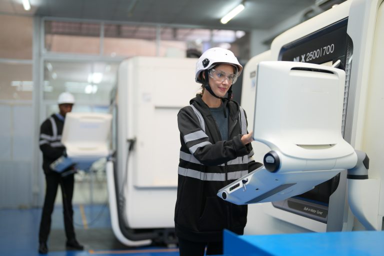 A female engineer stands at a computer screen to check on some robotic equipment. She is wearing PPE - clear safety glasses, a white hard-hat, and black overalls.