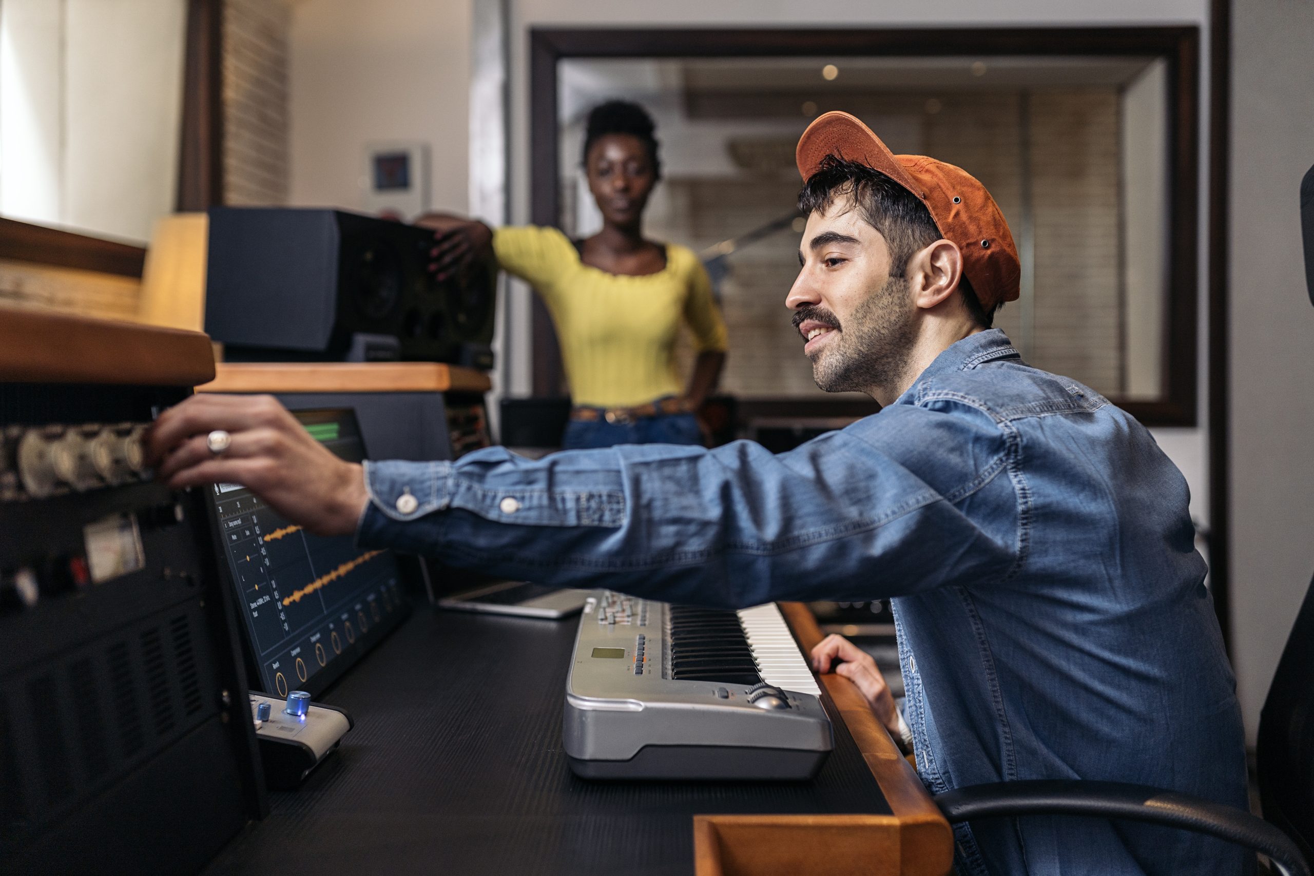 A man is working in a professional music studio. There is a woman standing in the background watching him work.