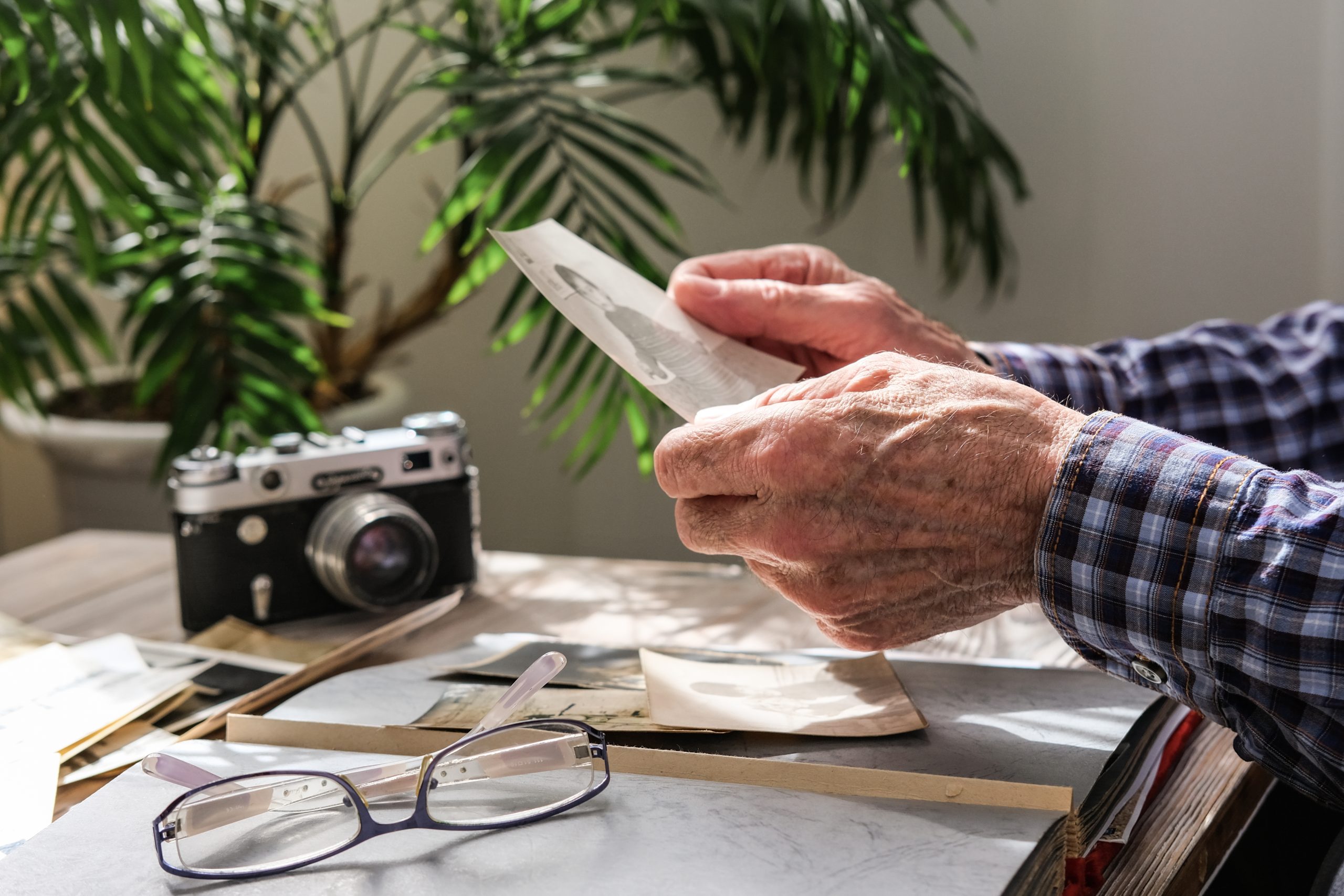 The hands of an elderly man hold a photograph. There's an old camera and a pair of glasses on the table