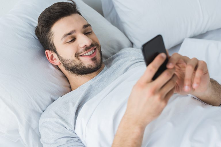Happy bearded man using smartphone in bed in the morning.