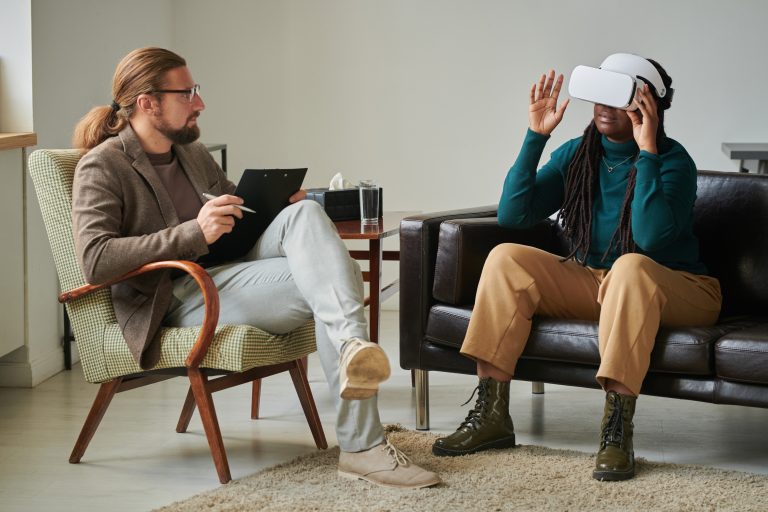 A man and a woman are sat in a therapy session. The woman is wearing wireless VR glasses as she speaks to the man. It looks like they are in therapy because he is taking notes.