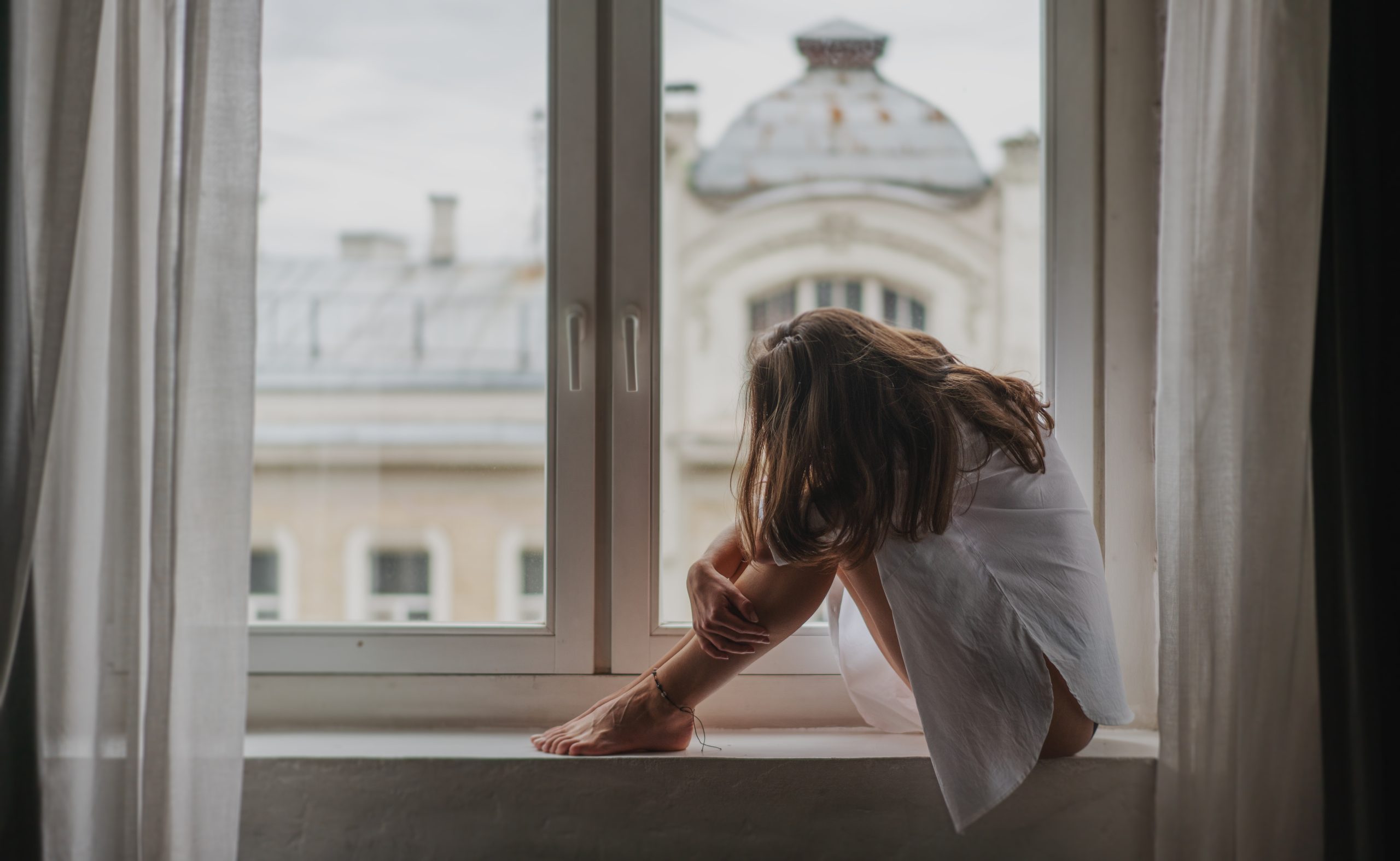 Young sad woman sitting on the windowsill by the window in the apartment hugging her knees and bowing her head