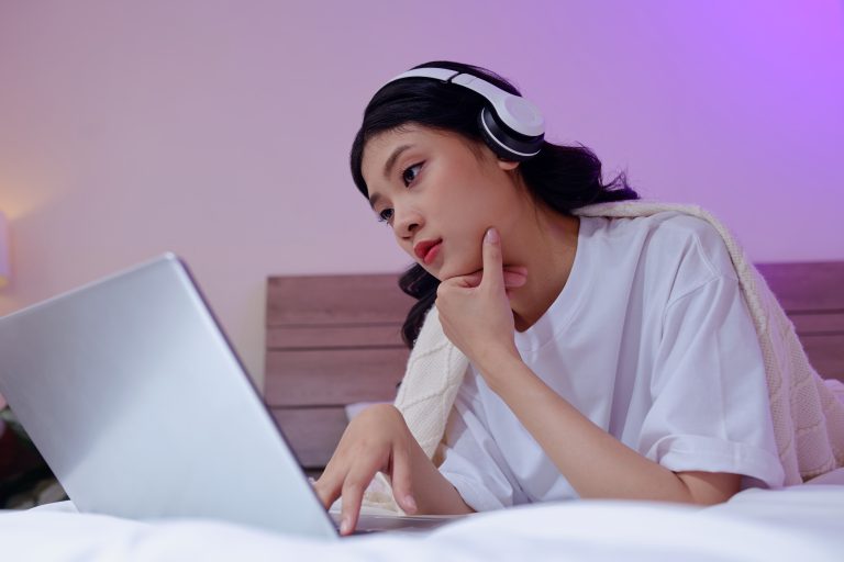 A woman lying on her bed with headphones on. She's looking contemplatively at her laptop.