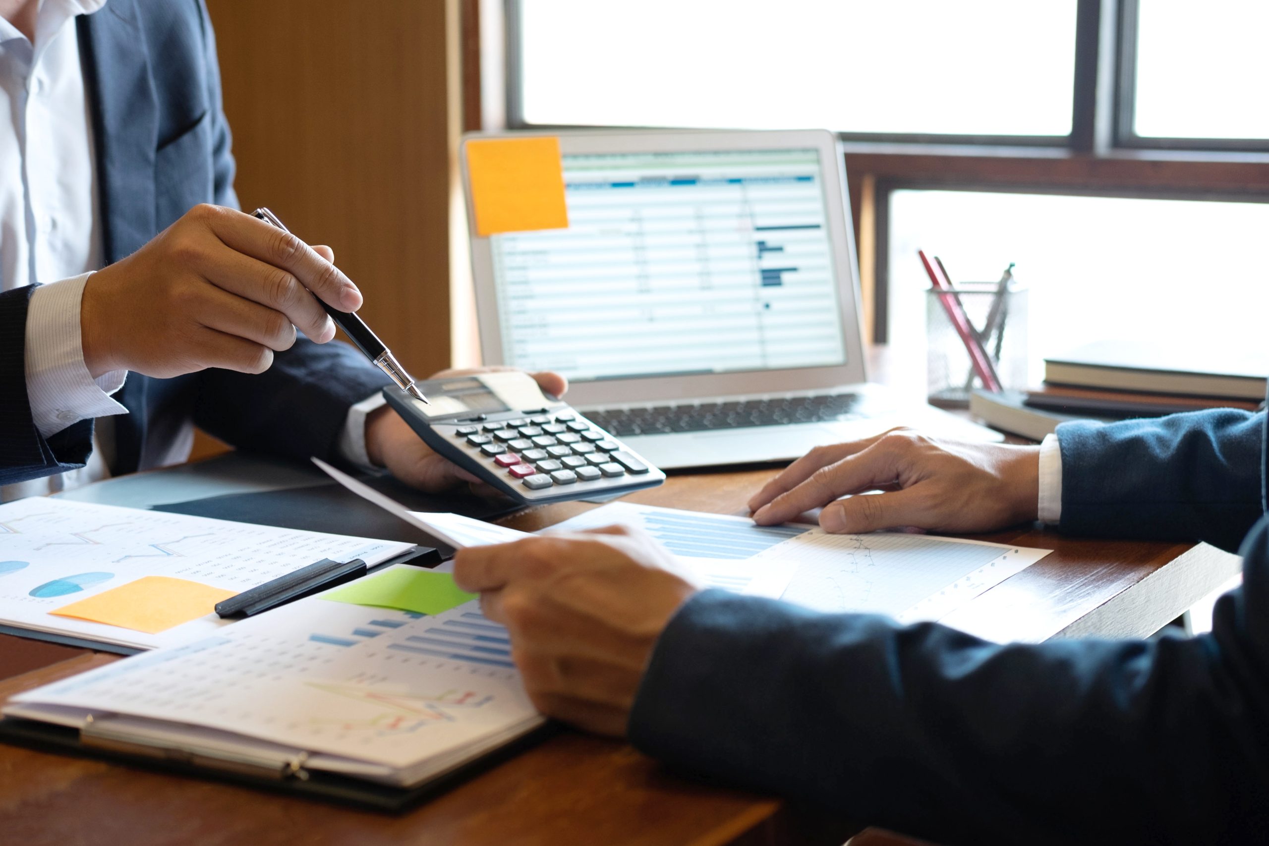 Two people sat opposite sides of a desk are having a business meeting. There are papers with graphs and calculators around them.