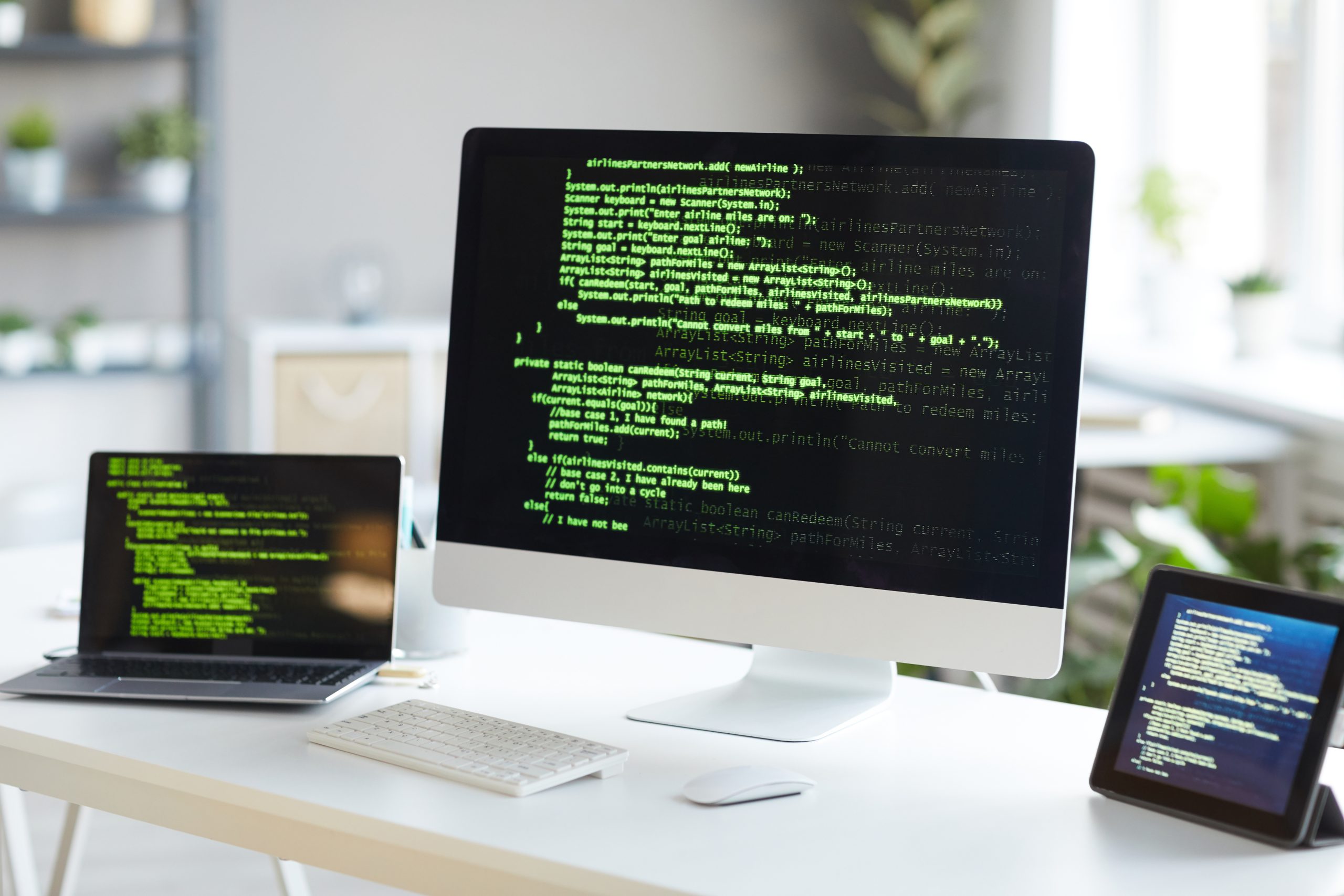 A compter, laptop, and tablet sit on a desk. Each one has green code and software on its screen.