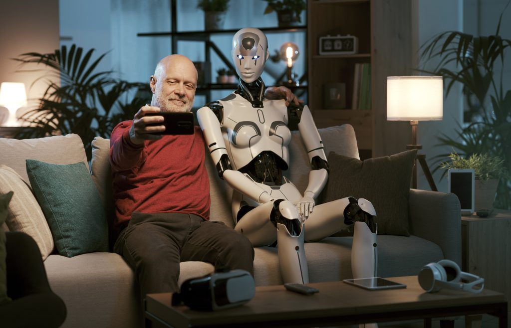An older man is sat on his couch at home. He is sitting next to a robot and they are taking a selfie together.