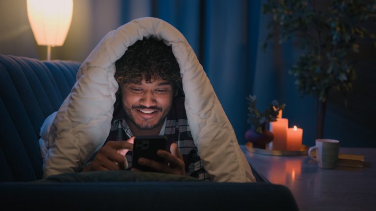 Smiling guy Indian man Arabian male at night evening home on couch under blanket cover with duvet looking mobile phone wow surprised amazed reading news gadget addict smartphone social media scrolling. High quality 4k footage