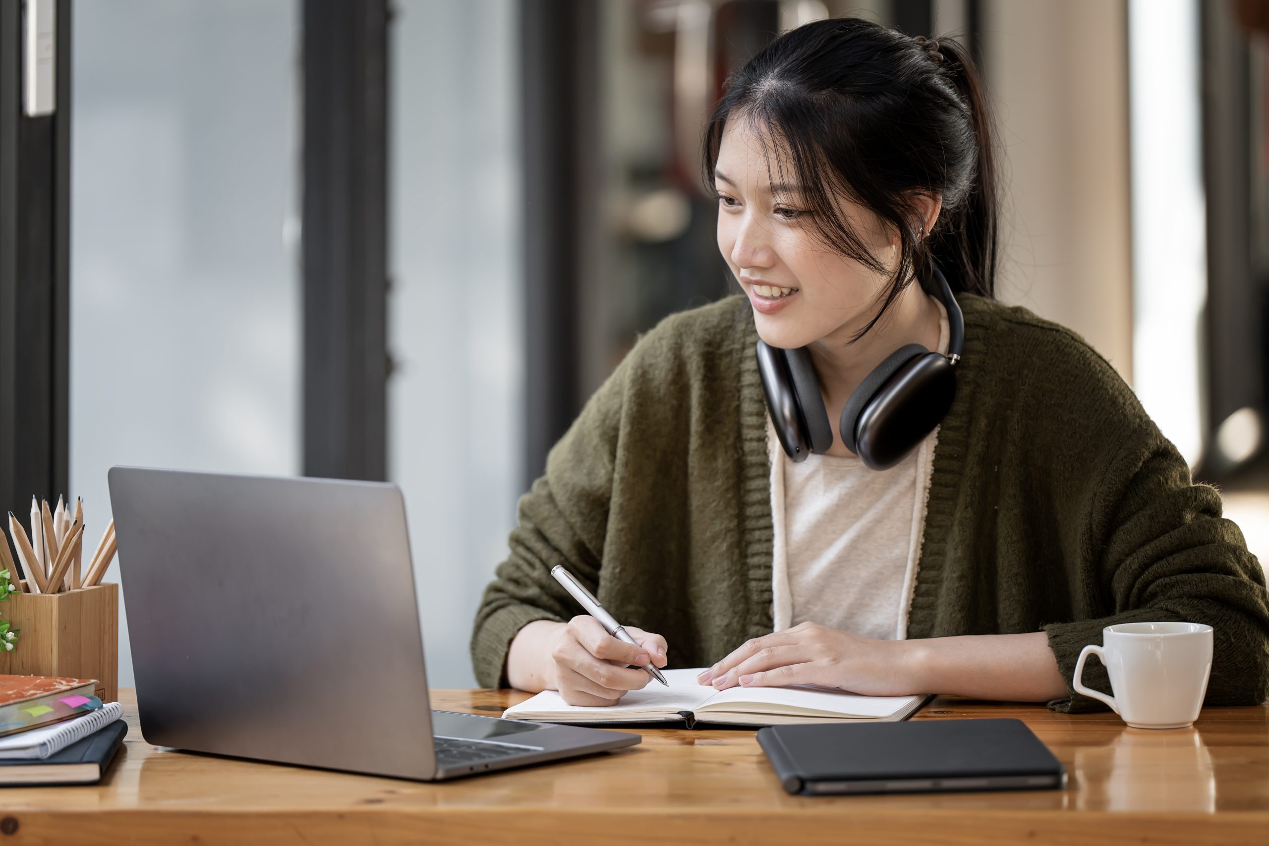 woman wearing headphones using laptop in cafe, writing notes, attractive female student learning online, listening audio course, e-learning education concept